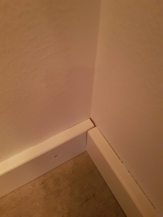 Vitmålade lister med synlig skruv och ojämn fogning i hörnet av ett rum.