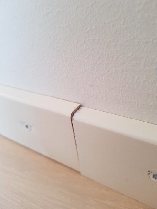 Vit golvlist med en synlig spricka vid hörnet och stora skruvar som är dåligt övermålade med vit färg.
