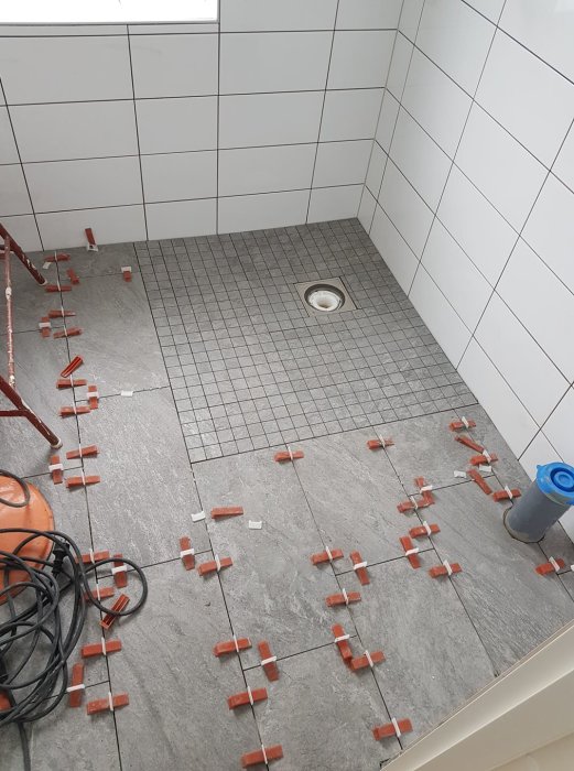 Badrum under renovering med nyinstallerad golvbrunn och gråa kakelplattor säkrade med röda kilar.