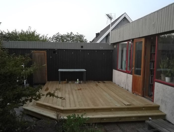 Träaltan ansluten till ett hus med svarta plank och röda fönsterkarmar, seedd från trädgården.
