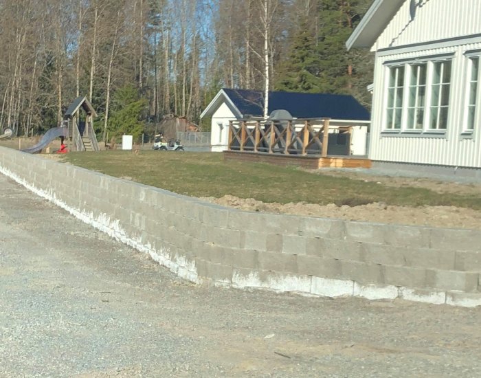 Nybyggd stödmur av gråa betongblock vid en trädgård nära ett hus och lekplats.