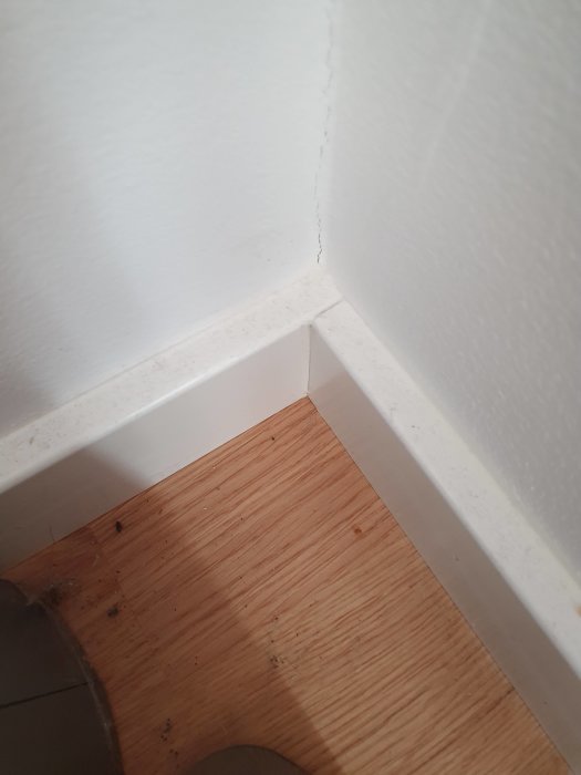 Innerhörn i ett rum där två raka vita lister möts utan gering på en trägolvbakgrund.