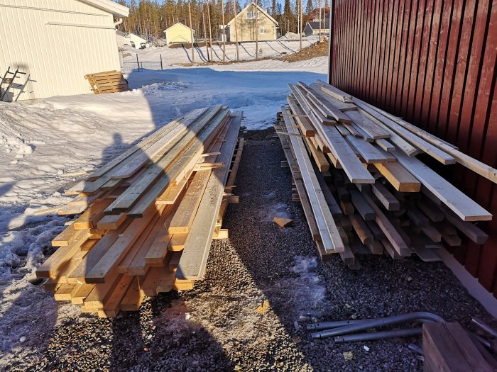 Staplade reglar och paneler förvarade utomhus bredvid en röd byggnad med snötäckt mark i bakgrunden.