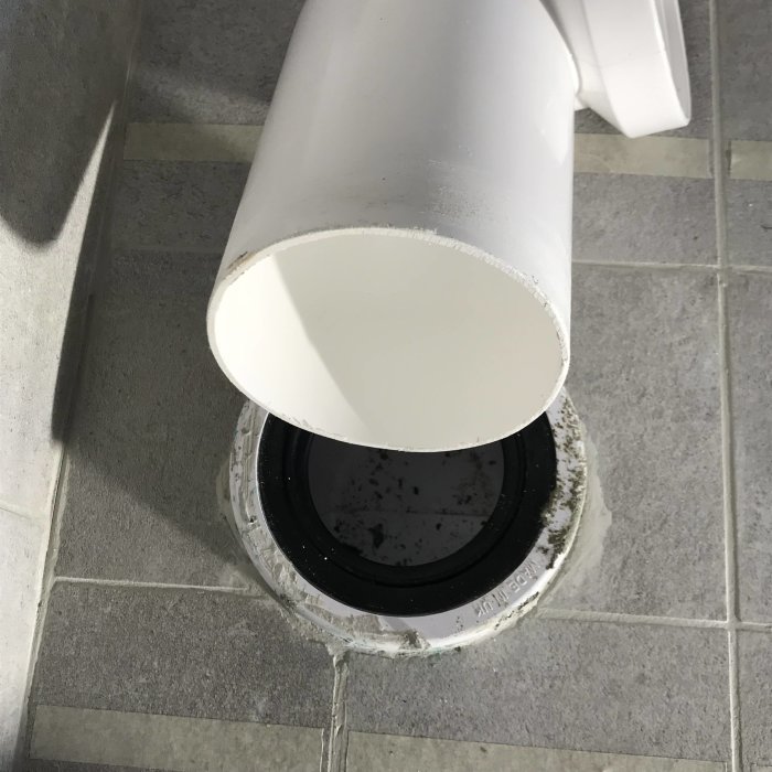 Vit klosettanslutning som inte är infogad i svart excenter i ett kaklat badrumsgolv.