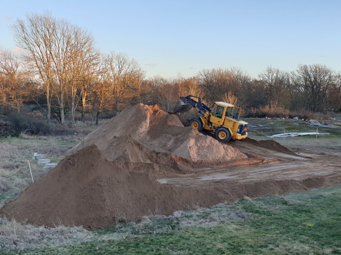 Gul hjullastare som tippa en hög med sand på en byggarbetsplats omgiven av träd.