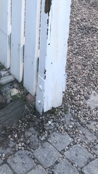 Gammalt vitmålat staket med slitet trä och befintliga metallfästen på en grusgång.