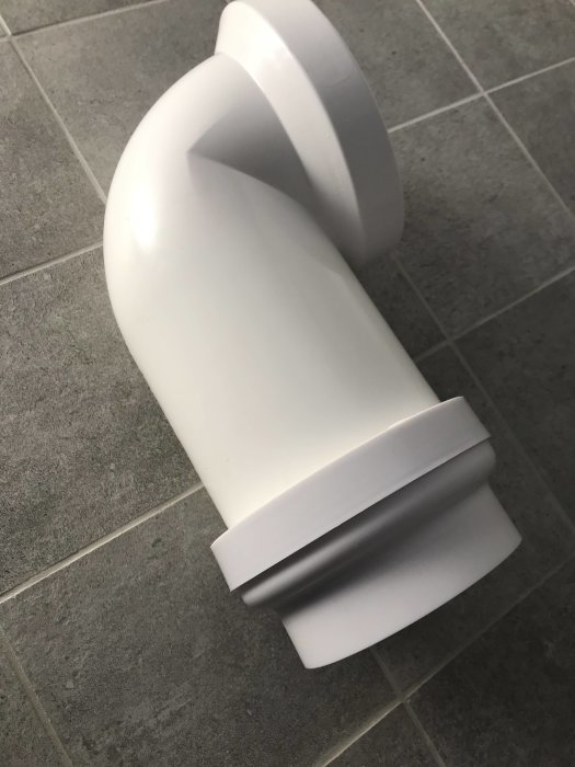 Vit plastadapter med böjd form på grått kakelgolv, hemmagjord lösning för rörinstallation.