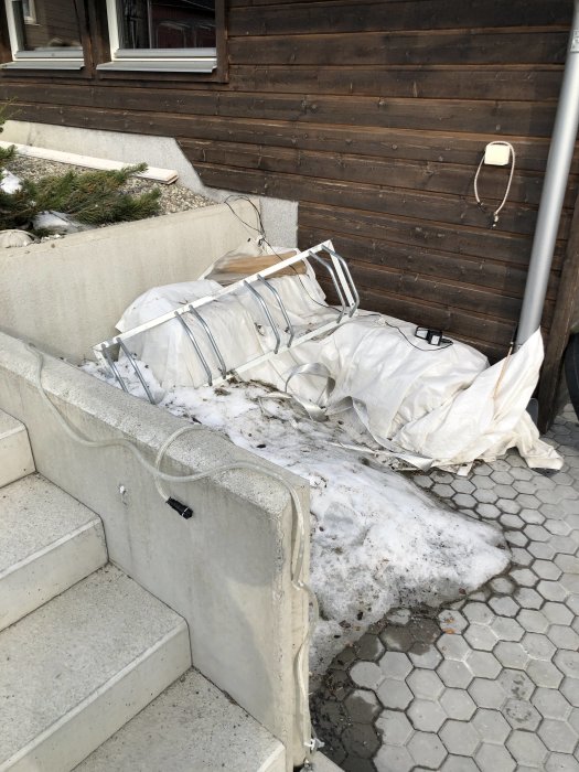 Hörna av hus med smältande snö och byggmaterial vid trävägg och stentrappa, planerat för förråd.
