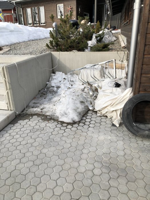 Hörna av en uteplats med snötäckt mark, betongplattor och husvägg, tänkt för ett förrådsbygge.
