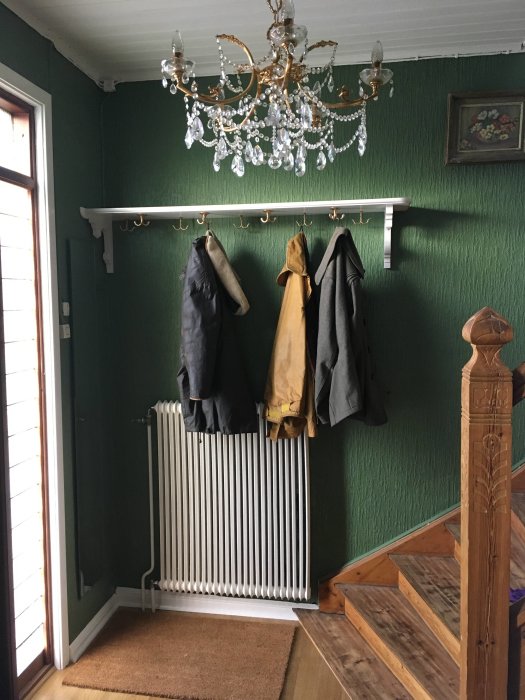 Vitmålad hatthylla med krokar och ytterkläder ovanför en radiator i en hall med grön tapet och kristallkrona.