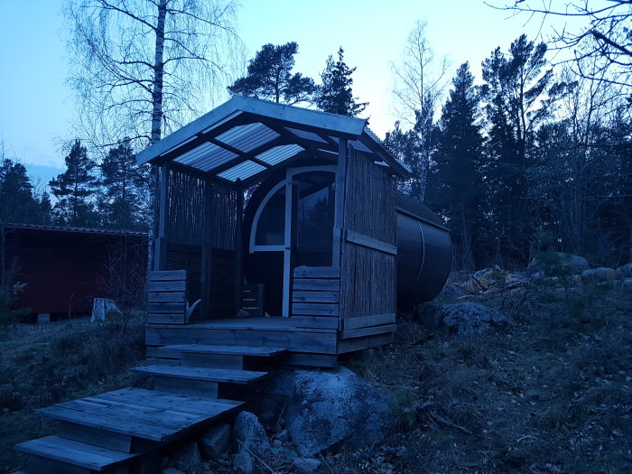 Ett småskaligt byggprojekt av en vedeldad bastu med stegar, omgivet av skog i skymningen.