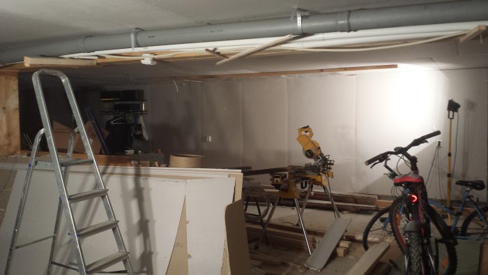 Garage under renovering med synliga rör i taket, en stege, såg och cyklar.