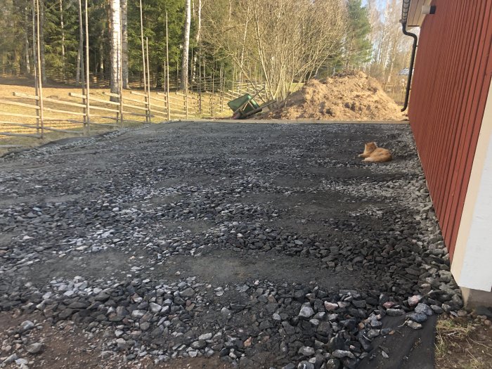 Anlagd parkeringsplats bredvid garage under arbete med bergskross och synlig lutning mot gärdesgård, katt sitter på gruset.