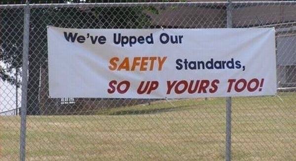 Banner hängande på staket med text "We've Upped Our SAFETY Standards, SO UP YOURS TOO!" som uppmanar till ökad säkerhetsmedvetenhet