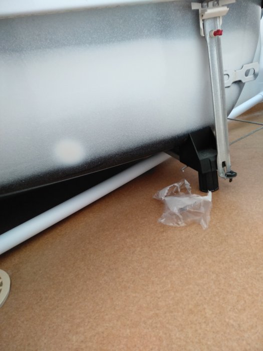 Vit PVC-rör ledning löst dragen under en radiator, ej fäst vid vägg, med öppet golv och plastförpackning.