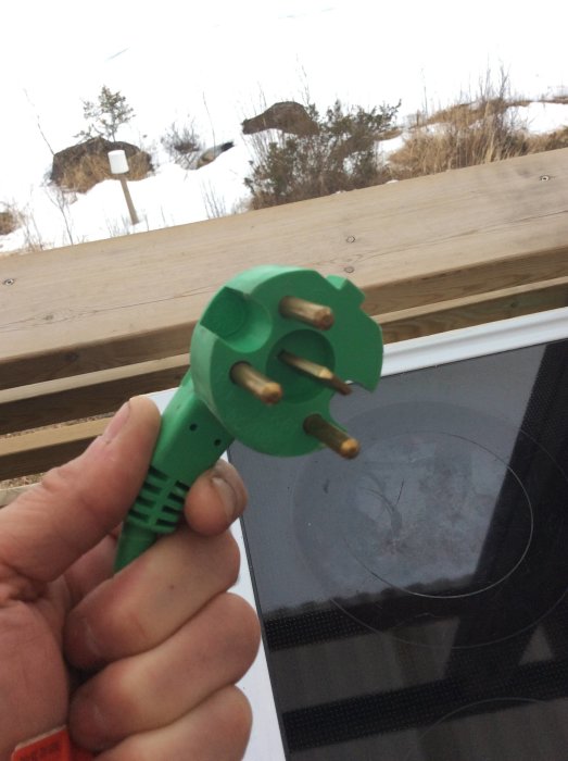Hand håller en grön fabriksmonterad elektrisk kontakt mot suddig utomhusbakgrund.