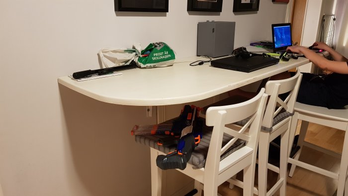 Hemmakontor med vitt skrivbord, laptop, spelkontroller, och en Nerf-blaster på en stol.