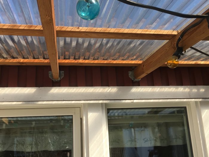 Altantak med träbjälkar och genomskinligt takmaterial över en husdel, med lodräta stödreglar och ljustaklampor.