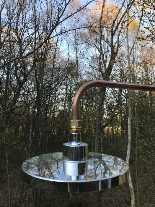 Närbild av en ny utedusch med kromad duschmunstycke mot en bakgrund av träd och himmel.
