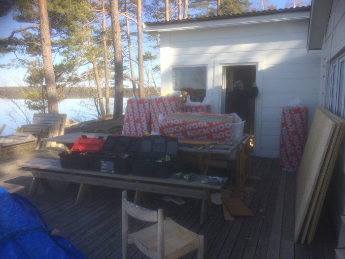 Byggmaterial och verktyg utspridda på träterrass vid hus med sjöutsikt, isolering i förgrunden, person i dörröppning.