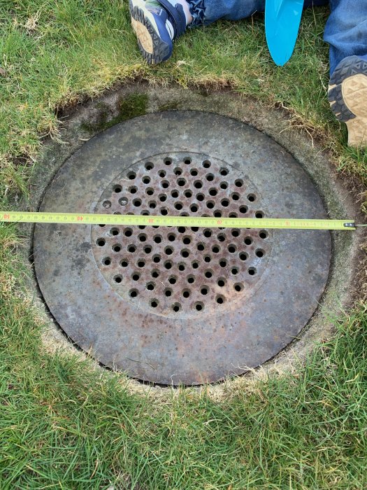 Rostigt cirkulärt lock med hålmönster över en stenkista, mätt med tumstock, i gräsmark med personens sko synlig.