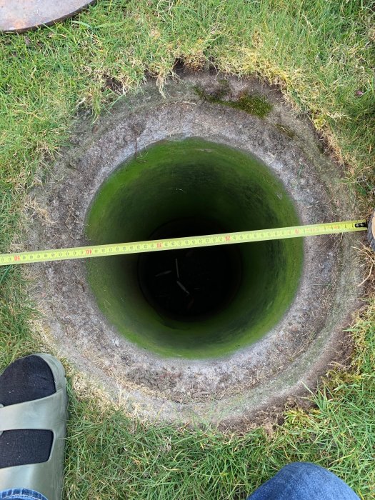 Öppen stenkista i trädgården med mätband över öppningen, intill grönt gräs och delvis synligt lock.