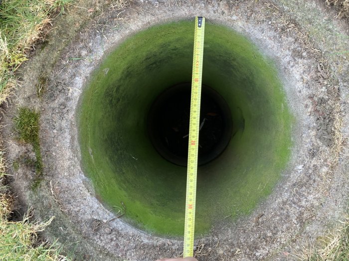 Måttband sträcker sig över öppningen av en grönaktig stenkista i trädgården, som mäter djupet av brunnen.