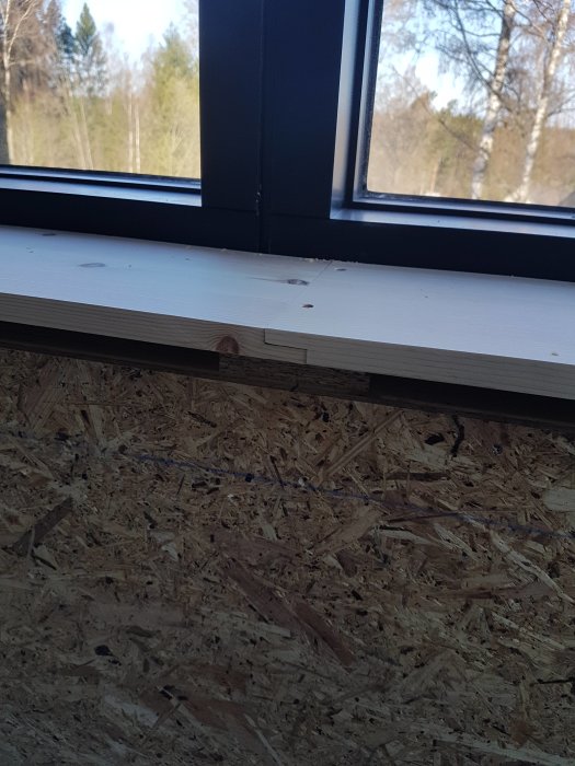 Nyinstallerat fönstersmyg vid svartmålat fönster med utsikt mot träd, ovanför OSB-skivor på vägg.