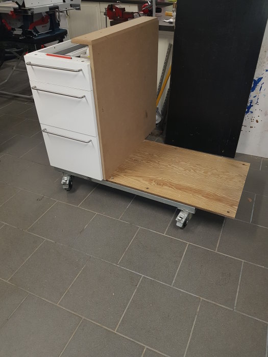 Byggprojekt med vit byrå monterad på en plyfa plattform med hjul i en verkstadsmiljö.