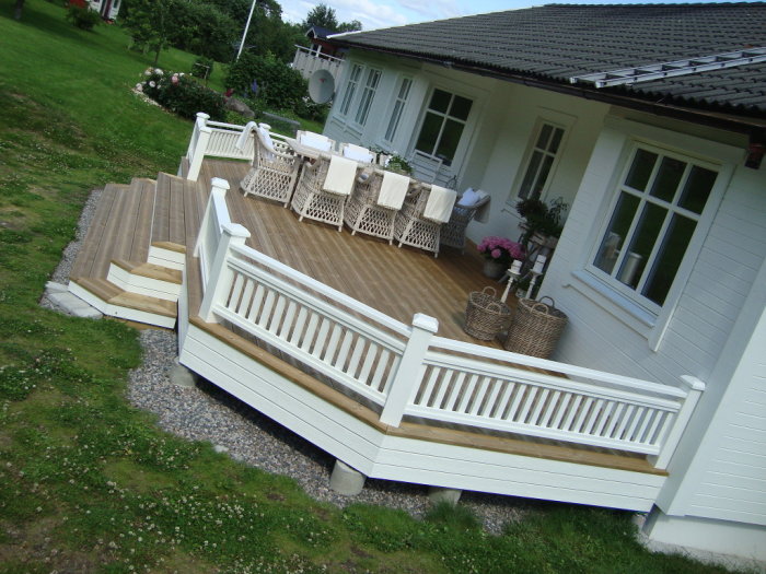 Altan med trädäck och vitt räcke, möblerat med bord och stolar, framför ett hus, anslutet till ytterdörren.