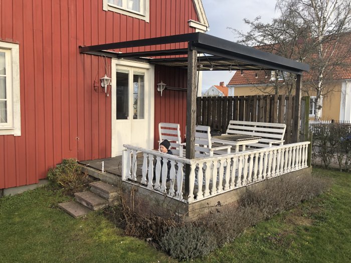 Röd trähusfasad med en vitmålad, ej färdigställd altan och pergola, överblivna betongplattor syns vid trappan.