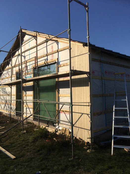 Delvis klädd fasad med träpanel och byggnadsställning runt ett hus under renovering.