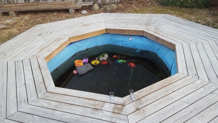 Oskött hexagonal hemmapool med smutsigt vatten och flytande leksaker omgiven av träaltan.