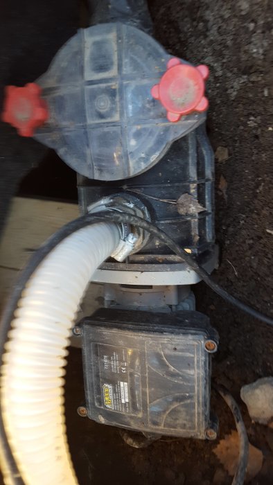 Smutsig poolpump och filter med anslutna slangar, placerad på marken bredvid en träbyggnad.
