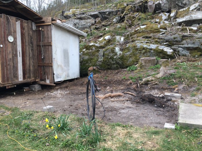 Rivet gårdshus där grunden och rötter syns, med en klippa och skog i bakgrunden.