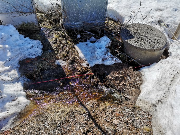 Brunn med öppet lock bredvid, delvis täckt av snö och omgiven av smältvatten.