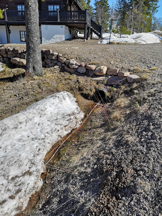 En friliggande kabel löper på marken med smältande snö nära ett stenlagt stödmur och en stuga i bakgrunden.