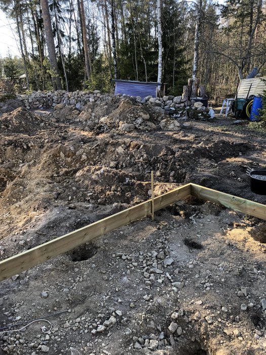 Uppgrävd trädgård med stenar och jord, påbörjad träram för altanbygge, bakgrunden visar skog och uthus.