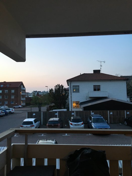 Vy från en loftgång med träplank och metallstaket med parkering och hus i bakgrunden under skymning.