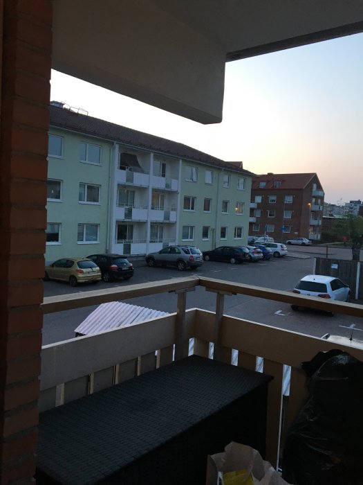Utsikt från loftgång med räcke vid flerfamiljshus under skymning, parkerade bilar syns.
