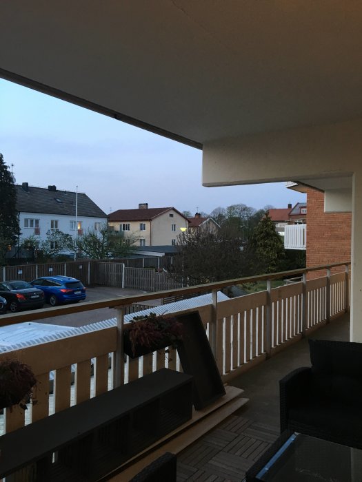 Utsikt från en lägenhetsdörr mot en loftgång med räcke och blomlådor, med hus och träd i bakgrunden.