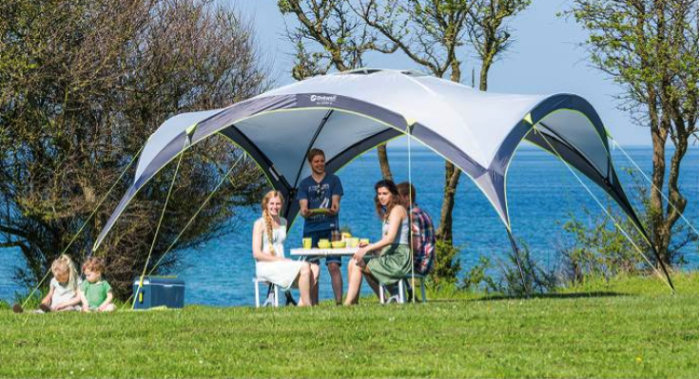 Familj under grått och vitt skyddstält Day Shelter från Outwell på en solig gräsmatta vid havet.