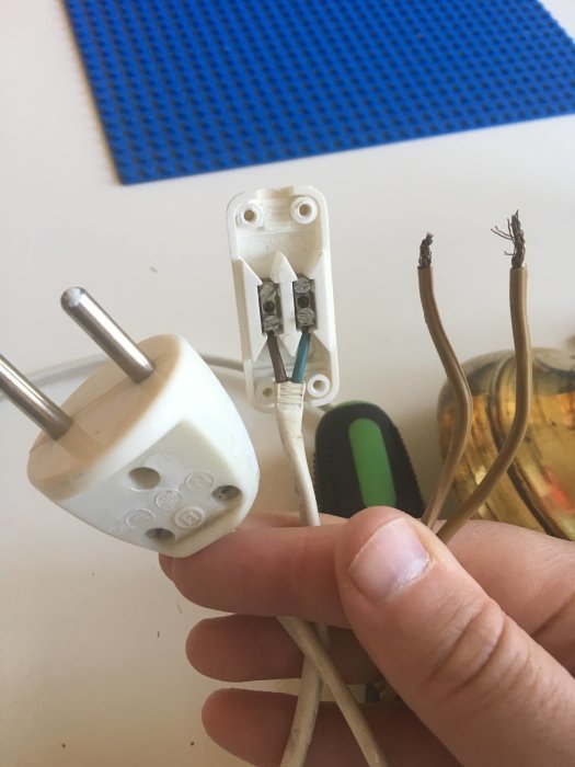 En hand håller en delvis isärskruvad vit stickkontakt med exponerade kablar i brunt och blått.