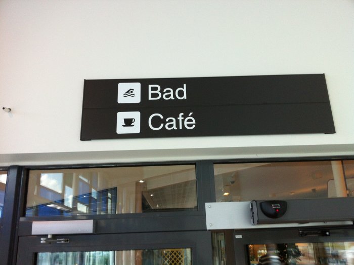 Skylt ovanför dörr med ikoner för "Bad" och "Café" i en simhall.