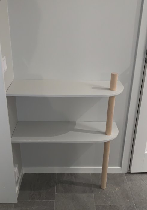 Egendesignat vitt sideboard med hyllor och en upprättstående träpinne i hörn, står på grått golv mot vit vägg.