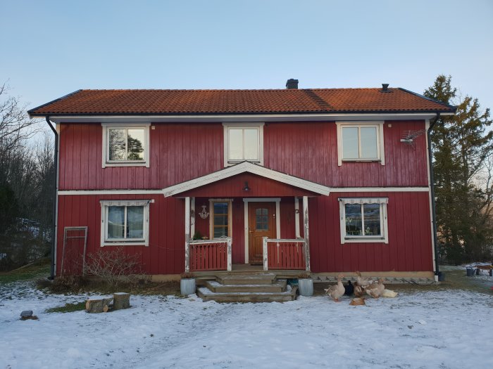 Tvåvåningshus från 1991 med röd stående panel och vita fönsterkarmar samt snö på marken.