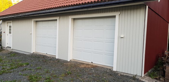 Garage med stående grå panel och vita dubbelgaragedörrar, kontrast mot röd sidopanel, hus i fokus för panelbyte.