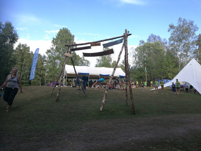 Scoutläger med barn och ledare vid tält och hemmagjord träkonstruktion under blå himmel.