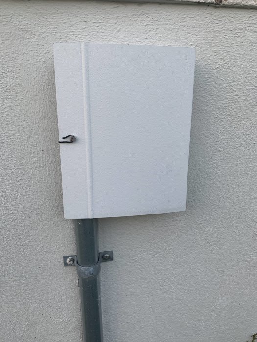 Stängd vit skarvbox på en husgrund med synlig grå kabel och rörinstallation på utsidan.