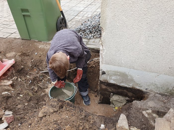 Person som lagar hål i marken vid husvägg, nära en pumpbrunn och kabelskyddsrör.